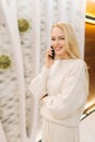 Vertical portrait of cheerful young woman talking on smartphone standing in hotel lounge, looking at camera. Cute blonde Royalty Free Stock Photo