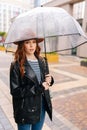 Vertical portrait of confident redhead young woman wearing elegant hat standing with transparent umbrella on street in Royalty Free Stock Photo