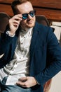 Vertical portrait of confident young man in sunglasses wearing stylish suit wear sitting in armchair, looking away out Royalty Free Stock Photo