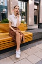 Vertical portrait of focused pretty young woman having smartphone conversation sitting on bench on city street in summer