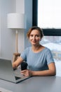 Vertical portrait of friendly blonde woman working on laptop computer, holding cup with coffee, looking at camera Royalty Free Stock Photo