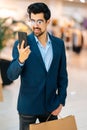 Vertical portrait of handsome bearded young man in fashionable suit and glasses using mobile phone standing in hall of Royalty Free Stock Photo