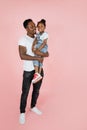 Vertical portrait of happy afro daddy embracing with his cute baby daughter, pink studio background. Royalty Free Stock Photo