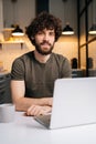 Vertical portrait of happy bearded young business man using typing on laptop sitting at table in kitchen room with Royalty Free Stock Photo