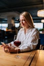 Vertical portrait of happy blonde young woman using smartphone, typing message sitting at table with glass of red wine Royalty Free Stock Photo
