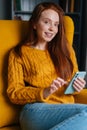 Vertical portrait of happy redhead young woman using smartphone sitting in yellow armchair smiling looking at camera. Royalty Free Stock Photo