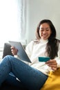 Vertical portrait of happy young Asian woman online shopping using digital tablet and credit card sitting on sofa. Royalty Free Stock Photo
