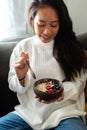 Vertical portrait of happy young Asian woman eating healthy smoothie breakfast bowl of oats and fruit on the couch. Royalty Free Stock Photo