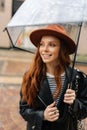 Vertical portrait of joyful ginger young woman wearing elegant hat standing with transparent umbrella on city street Royalty Free Stock Photo