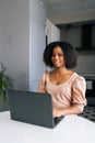 Vertical portrait of positive black female freelancer sitting at table in modern kitchen room working on laptop, smiling Royalty Free Stock Photo