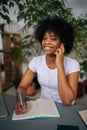 Vertical portrait of positive black female student in headphones holding pen in hand sitting at desk with laptop, paper Royalty Free Stock Photo