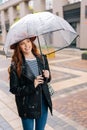 Vertical portrait of positive young woman in stylish hat standing with transparent umbrella on city street enjoying Royalty Free Stock Photo