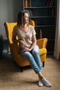 Vertical portrait of pretty blonde woman sitting in yellow chair in dark room with modern interior, thoughtful looking Royalty Free Stock Photo