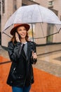 Vertical portrait of pretty ginger young woman wearing elegant hat standing with transparent umbrella talking on mobile Royalty Free Stock Photo