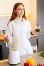 Vertical portrait of pretty redhead young woman holding glass with green vegetable detox smoothie cocktail from blender Royalty Free Stock Photo