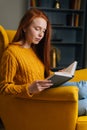 Vertical portrait of pretty young woman reading exciting story having relaxing weekend sitting in yellow armchair. Royalty Free Stock Photo