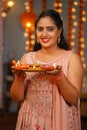 Vertical portrait shot of Happy Indian young woman looking at camera by holding diwali diya lamp at home - concept of