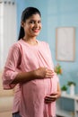 vertical Portrait shot of happy smiling Indian Pregnant woman standing by holding tummy by looking at camera - concept