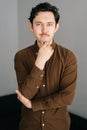 Vertical portrait of smiling young man with little moustache holding hand on chin and thoughtful looking at camera Royalty Free Stock Photo