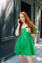 Vertical portrait of smiling young woman in green dress holding takeaway coffee cup, talking on mobile phone walking at Royalty Free Stock Photo