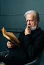 Vertical portrait of thinking mature gray-haired aged male reading paper book sitting on chair in dark living room Royalty Free Stock Photo