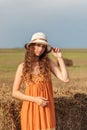 Vertical portrait of a young cute rural woman in an orange sarafan and a hat near a stack of wheat hay on a field in the rays of t