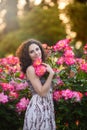 A vertical portrait of young Caucasian woman with dark brown curly hair near pink rose bushes, looking to her left, smiling with Royalty Free Stock Photo
