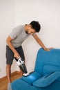 Vertical portrait of young man using handheld vacuum cleaner to remove dust and dirt from blue sofa, emphasizing Royalty Free Stock Photo