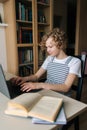 Vertical shot of adorable little schoolgirl using typing on laptop keyboard sitting at table with paper textbook Royalty Free Stock Photo