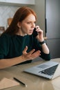 Vertical shot of angry young woman talking on mobile phone and using laptop sitting at table in kitchen with modern Royalty Free Stock Photo