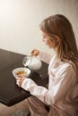 Vertical shot of attractive blonde daughter sitting in cozy pyjamas in kitchen, pouring milk in bowl with cereals Royalty Free Stock Photo