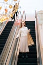 Vertical shot of attractive young woman shopper holding on escalator handrail and riding escalator going down in Royalty Free Stock Photo