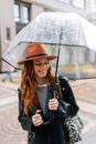 Vertical shot of cheerful young woman in hat standing with transparent umbrella on city street enjoying rainy weather Royalty Free Stock Photo