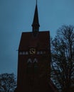 Vertical shot of a church tower with a clock and silhouetted trees against a blue evening sky Royalty Free Stock Photo