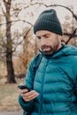 Vertical shot of concentrated hipster guy with thick beard, wears anorak and hat, holds cellular, uses special application for