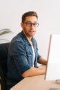 Vertical shot of confident young frelancer male in stylish glasses working on desktop computer sitting at desk at home Royalty Free Stock Photo