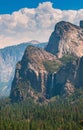 Vertical shot of forested mountains with a waterfall under a blue cloudy sky at daytime Royalty Free Stock Photo