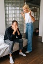 Vertical shot of frustrated Caucasian husband sitting on bed ignoring angry standing blonde wife arguing blaming upset