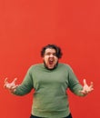 Vertical shot of a furious overweight man with a curly hair, standing in front of the red wall, feeling angry and frustrated, Royalty Free Stock Photo