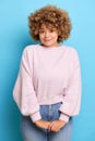 Vertical shot of gentle young woman with curly hair keeps arms together looks shy wears casual fluffy sweater and jeans Royalty Free Stock Photo