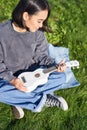 Vertical shot of girl musician, looking with care at her white ukulele guitar, playing music in park, sitting on grass Royalty Free Stock Photo