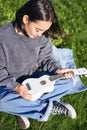 Vertical shot of girl musician, looking with care at her white ukulele guitar, playing music in park, sitting on grass Royalty Free Stock Photo