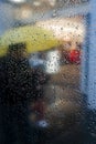 Vertical shot of a glass with raindrops and a person with a yellow umbrella in the background
