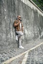 Vertical shot of good looking bold bearded guy standing outdoors against grey wall with cell phone in his hand chatting and Royalty Free Stock Photo