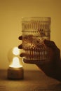 Vertical shot of a hand holding a patterned glass next to a lamp, artsy aesthetic