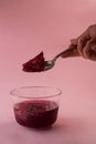 Vertical shot of a hand with a spoon picking up purple jelly from a bowl on a pink background