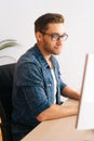 Vertical shot of handsome young designer male in glasses working on desktop computer sitting at desk at home office in Royalty Free Stock Photo