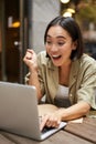 Vertical shot of happy girl talking on video call, looks at laptop, having online meeting, sitting in outdoor cafe Royalty Free Stock Photo