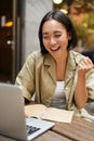 Vertical shot of happy girl talking on video call, looks at laptop, having online meeting, sitting in outdoor cafe Royalty Free Stock Photo
