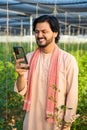 vertical shot of Happy smiling young farmer busy using mobile phone at greenhouse - concept of technology, modern